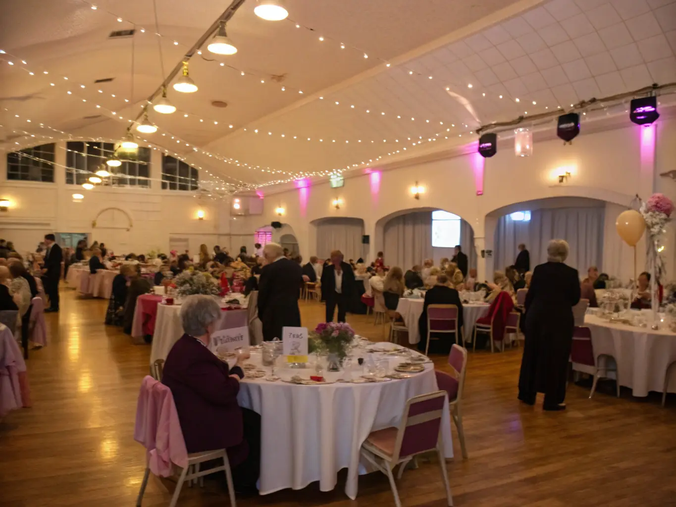 A dynamic image of a large hall decorated for a charity gala, with elegantly dressed guests, auction tables, and a stage where a speaker addresses the audience, capturing the excitement of a fundraising event.